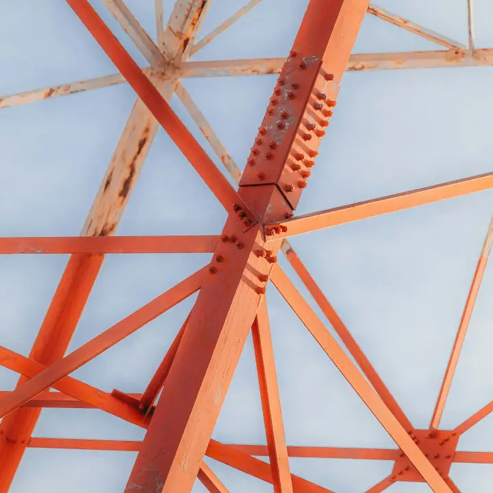Treillis d'acier orange vu d'en dessous, triangles boulonnés sur ciel pâle