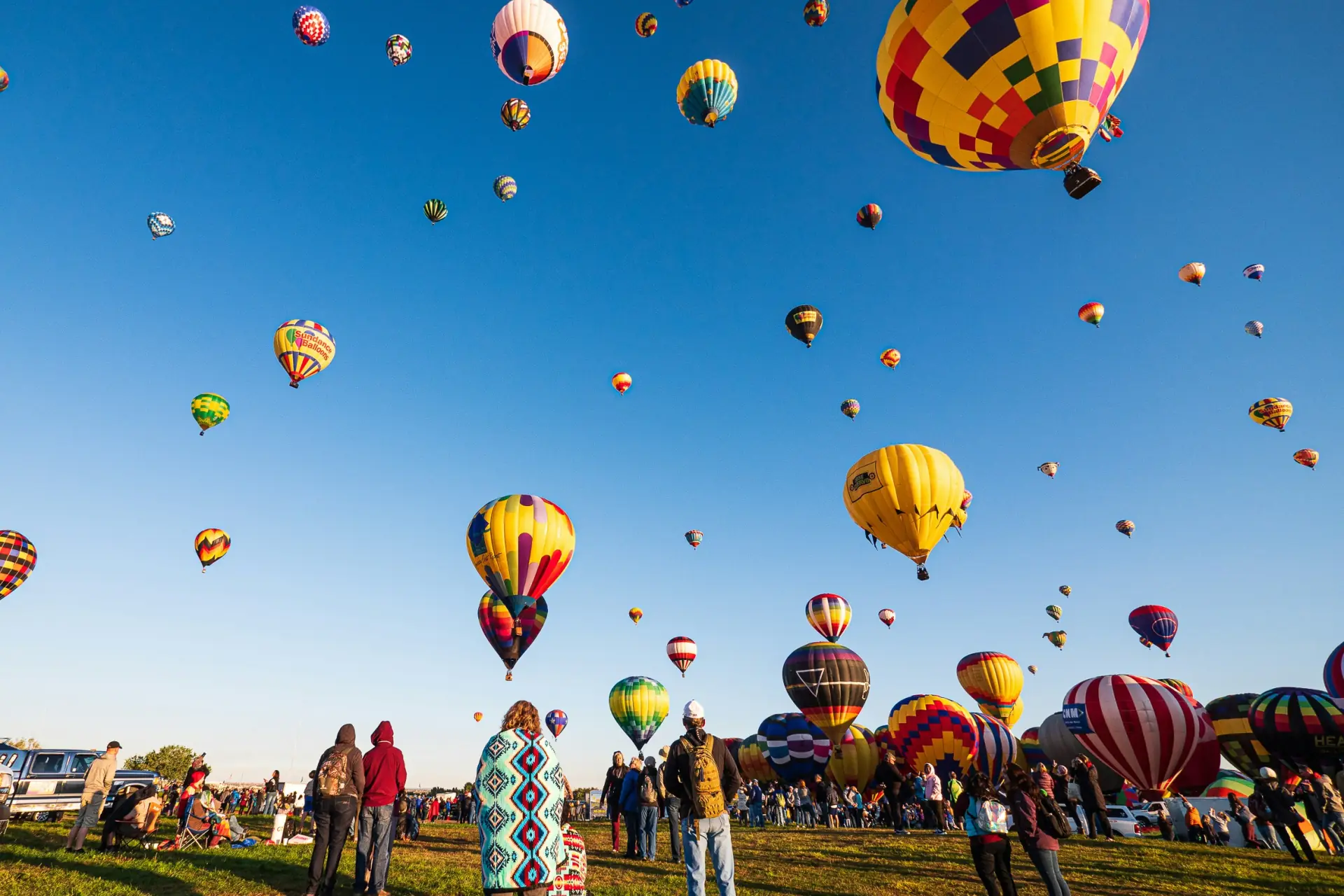 Foule vue de dos dans un champ, ciel bleu rempli de montgolfières multicolores