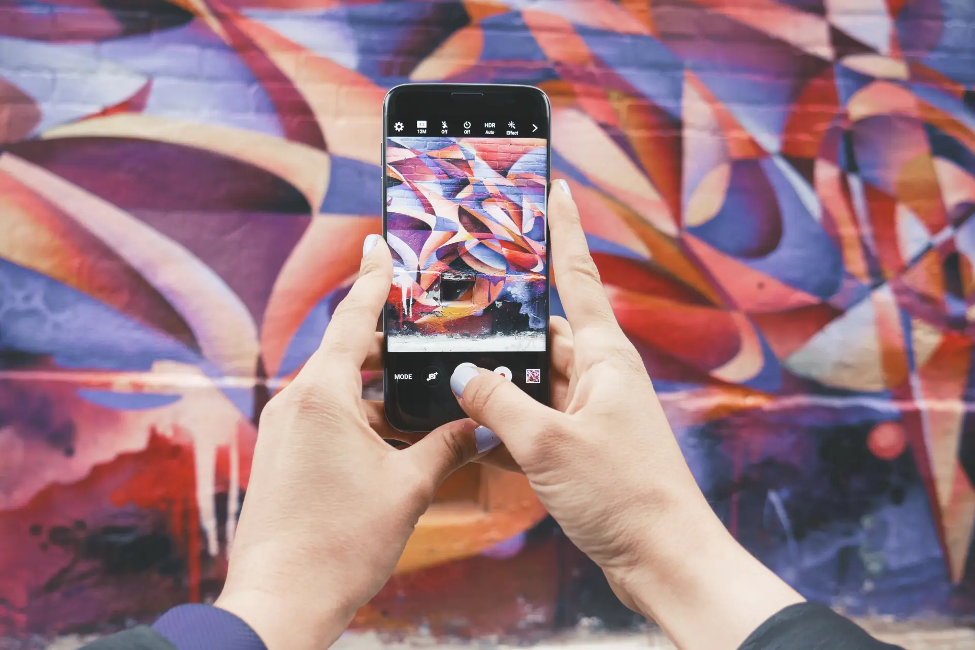 Mains tenant un téléphone devant une murale géométrique aux tons rouges et violets